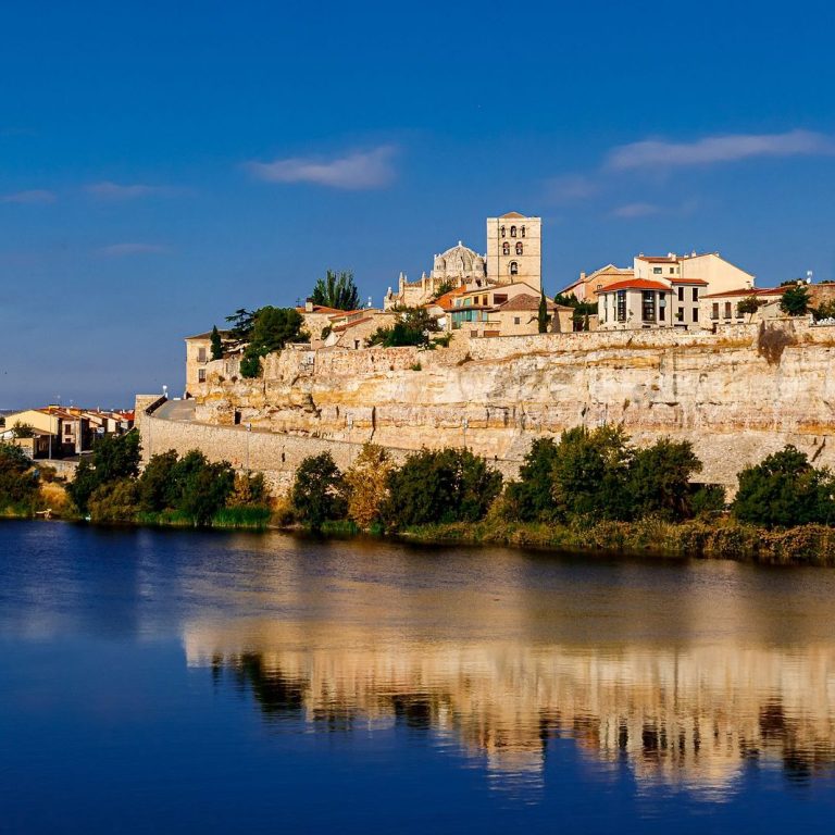 Vista de un pueblo en una colina junto a un río, con casas y un campanario.