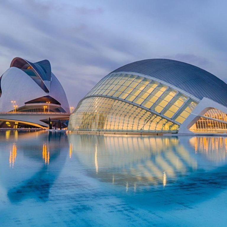 Edificios modernos en Valencia reflejados en una superficie de agua tranquila al atardecer.