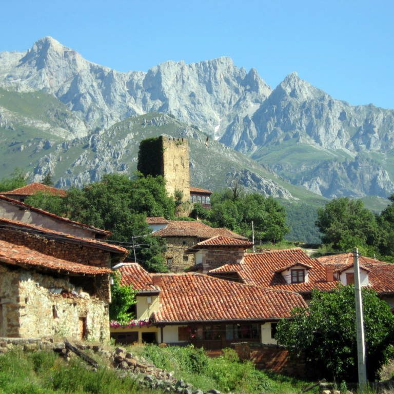 Vista de un pueblo con casas de piedra y un torreón, rodeado de montañas.
