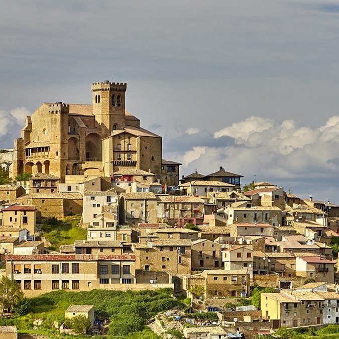 Castillo sobre colina junto a un pueblo con casas de varios colores y nubes en el cielo.