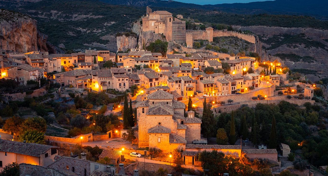 Vista nocturna de un pueblo medieval iluminado con luces cálidas de huesca