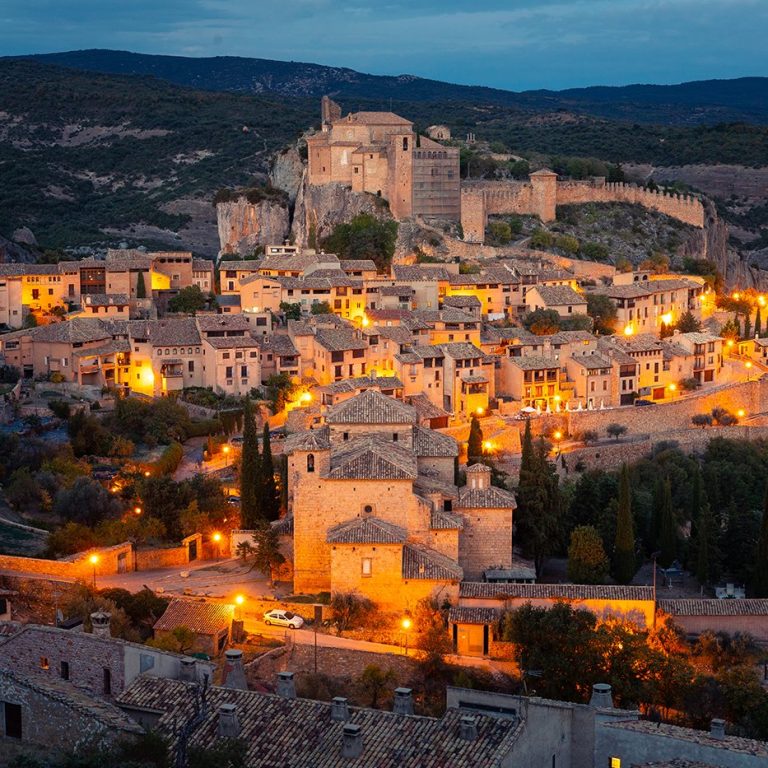 Vista nocturna de un pueblo medieval iluminado, con un castillo en la cima de la colina.