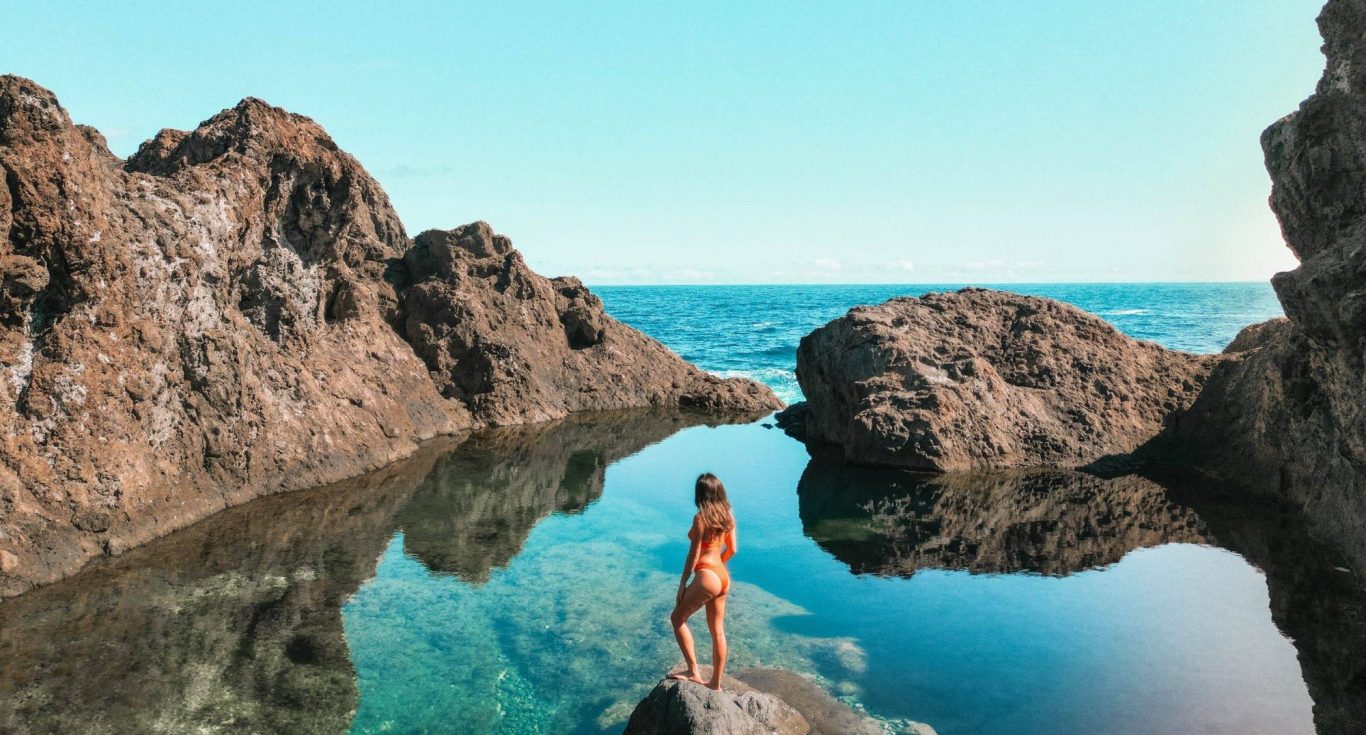Una persona de pie junto a un charco, con rocas y mar al fondo bajo un cielo azul de Tenerife