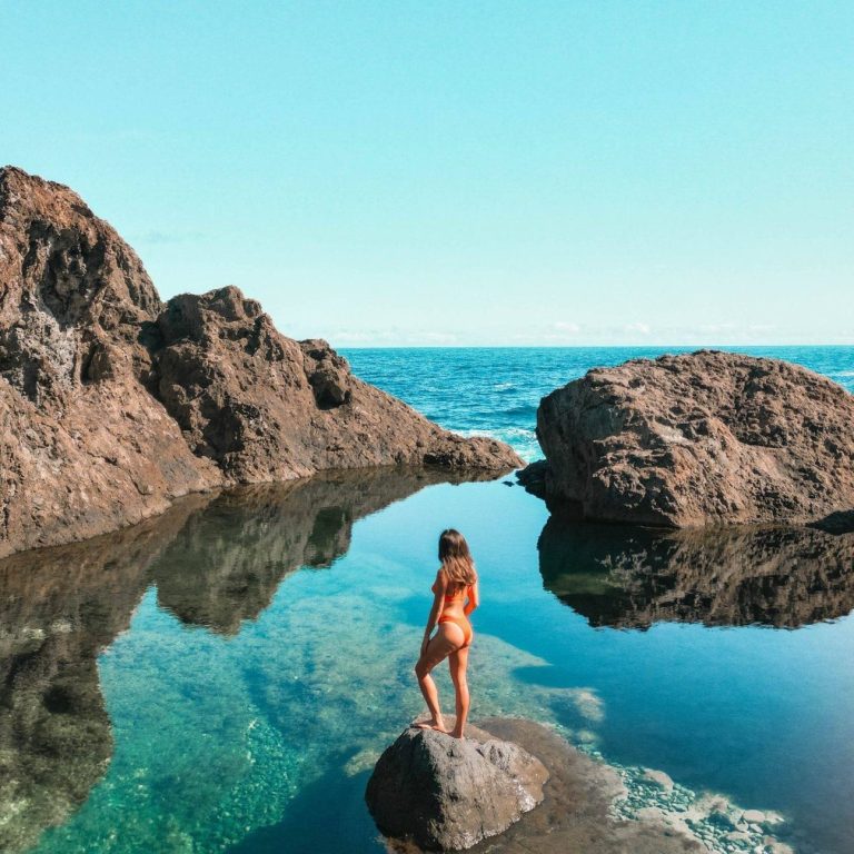 Niña de pie sobre una roca, contemplando el mar y reflejándose en el agua.