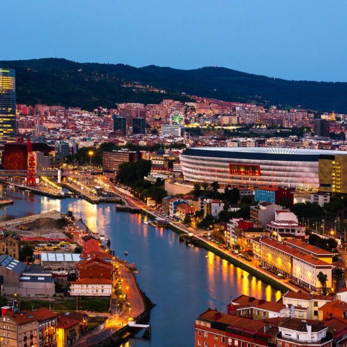 Vista nocturna de Bilbao con el río, edificios iluminados y colinas al fondo.
