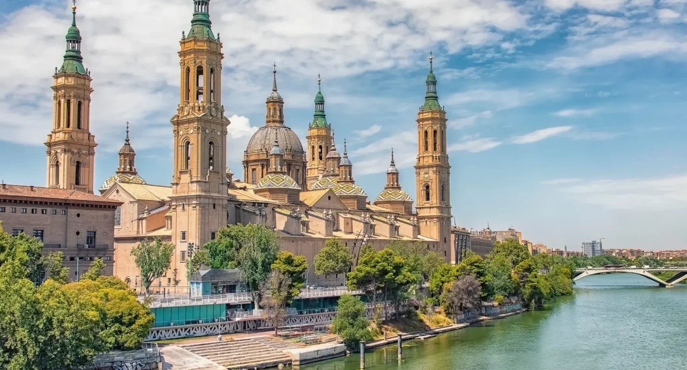 Basílica del Pilar junto al río Ebro, con cielos azules y vegetación circundante en Zaragoza
