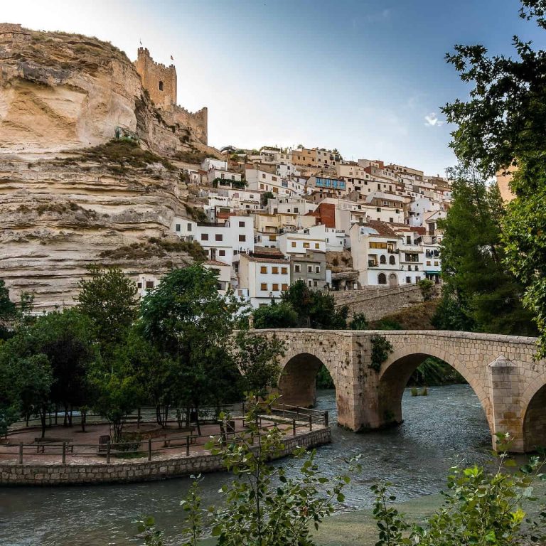 Puente de piedra sobre el río con paisajes montañosos y un castillo al fondo.