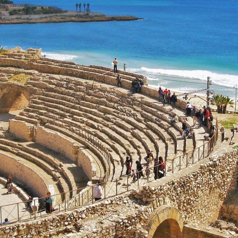 Ruinas de un anfiteatro antiguo junto al mar, con visitantes explorando su estructura.